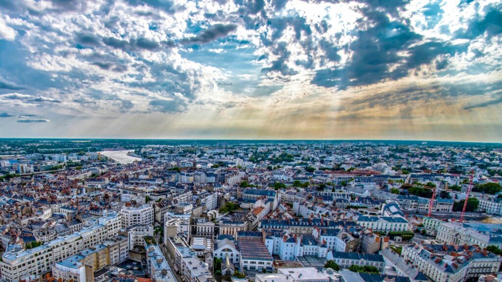 Vue aérienne de la ville de Nantes sous un ciel nuageux avec des rayons de soleil filtrant à travers les nuages.